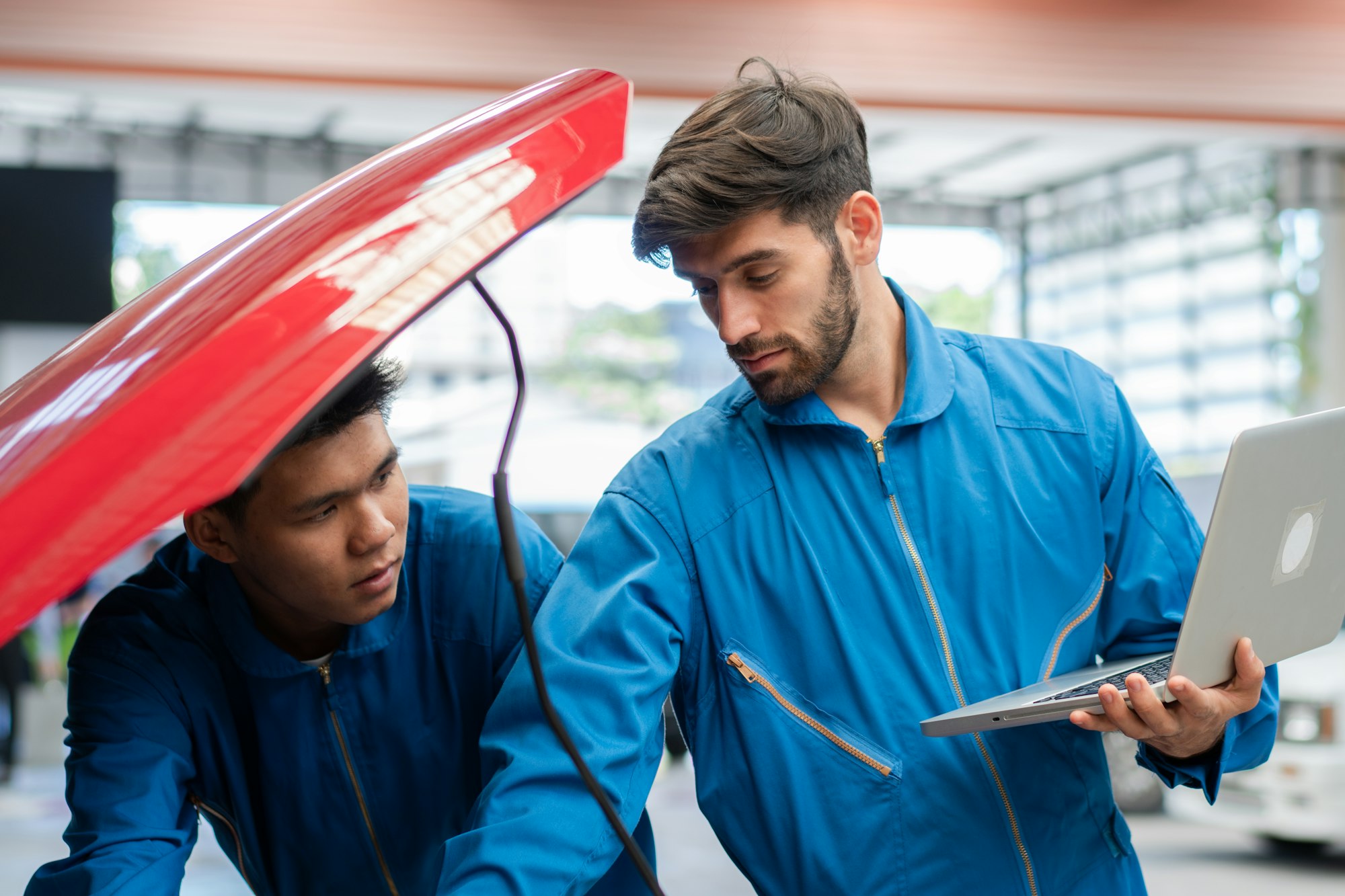 Mécanicien automobile caucasien utilisant un ordinateur portable pour diagnostiquer et réparer une voiture pendant que son collègue vérifie le capot du radiateur dans un garage automobile, entretien d'un technicien automobile après l'entretien